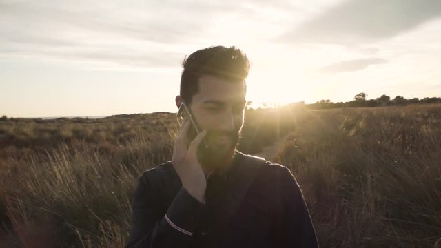 Content adult man with beard in stylish outfit speaking on phone against countryside in sunlight.
