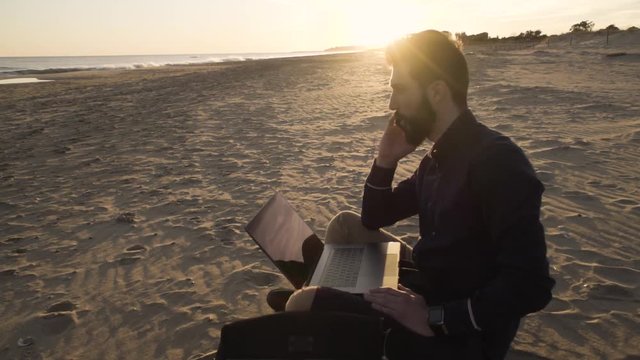 Concentrated man relaxing on a beach browsing laptop in sunlight.