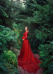 An elegant, pregnant woman walks in a beautiful garden in a luxurious, expensive red dress with a...