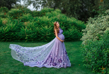 An elegant, pregnant woman walks in a beautiful garden in a luxurious, floral purple dress with a...