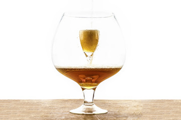 Beer is pouring into a glass which is into another huge glass, creating foam and bubbles on the wooden texture  at the bottom and white background