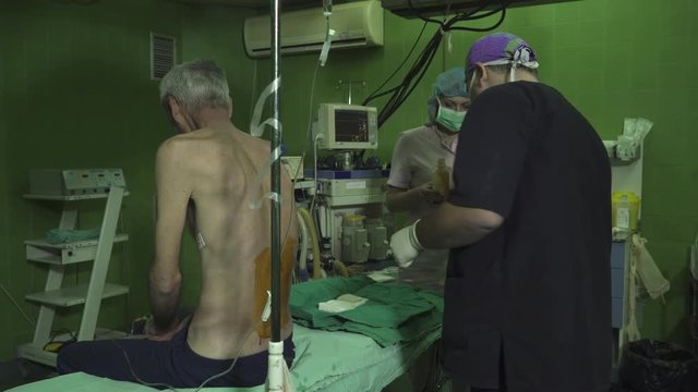 Mature, skinny patient sitting on surgical table, anesthesiologist using sterile gauze and scissors to disinfect back with iodine solution to injecting anesthetic, surgery operation in operating room