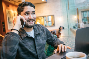 Happy phone chatting man in cafe with computer. Freelancer employed out office workplace. Big city lifestyle