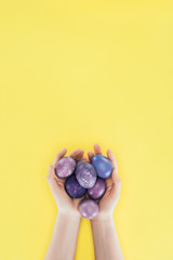 Cropped view of woman holding purple easter eggs in hands, isolated on yellow