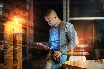 Concentrated reading. Calm attentive young man looking concentrated while standing next to the table with a bottle of beer in his hand and reading an article on the screen of his convenient tablet