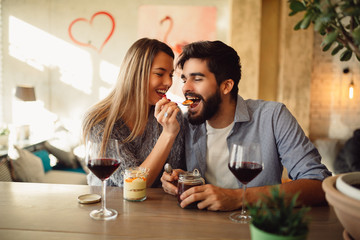 Beautiful couple in love is sitting in cafe, drinking red wine and eating cake. Young woman is feeding her man. Looking softly on each other.