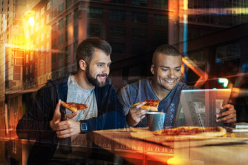 Eating together. Cheerful friendly young people smiling and looking at the screen of a modern laptop and relaxing while eating pizza