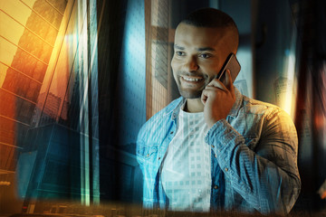 Phone talk. Positive friendly young man looking glad while standing near the window and having a...