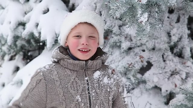 Cute little boy having fun outdoors in city street near snowy branches of huge old evergreen pine. Portrait of funny child blowing air kiss into camera. Slow motion.