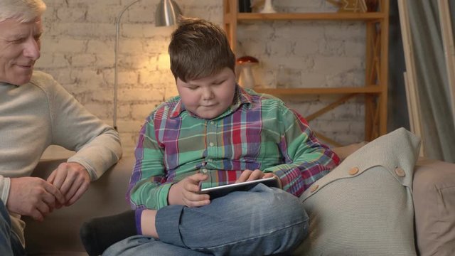 Young fat boy is sitting with his grandfather on the couch and playing on the tablet. The difference of generations. Home comfort, family idyll, cosiness concept. Clode up 60 fps