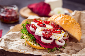 Vegitarian burger with beet slices, microgreen sprouts, tofu cheese, dried tomatoes and onion on kraft package paper on dark stone background with inhrediants. Soft selective focus. Copy space.