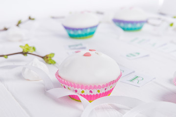 Fototapeta na wymiar Decorated Easter cupcakes on the white wooden table with branches with young shoots of greenery, satin ribbon and light merengue sweets. Holiday concept. Selective focus, copy space.