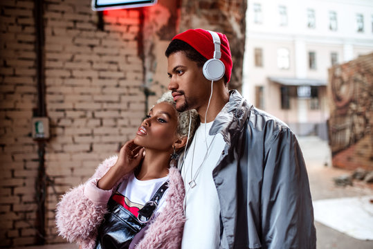 Attractive woman and her dark skinned stylish friend who listens music with headphones, walk together in street, looks at something with thoughtful expression. People and lifestyle concept.