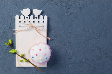 Simple Easter holiday composition of decorated cupcake, branch with young shoots of greenery and handcraft notebook with empy blank on dark stone background. Selective focus. Space for text.