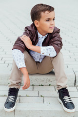Adorable little boy sitting on stairs in a city, wearing a brown leather jacket.