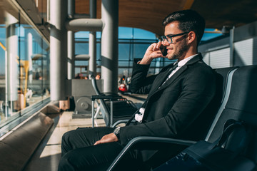 Young business man sitting on the phone with the suitcase at the airport waiting for the flight