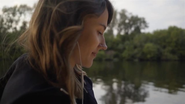 Side view of a young woman wearing a black hoodie listening to the music and walking along a river bank. Tracking real time close up shot
