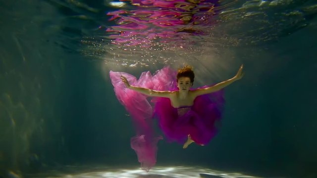 young woman with long red hair under water like in a fairy tale floats in a dress the color fuchsia
