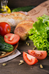 Fresh ingredients for cooking salad: tomato, cucumber, lettuce, olive oil and spices over rustic wooden table background. Served with fresh baguette. Healthy eating concept