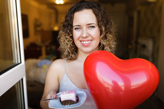 Beautiful curly young woman holding a dessert on a plate and a heart shape balloon. Valentine's Day concept, symbol of love.