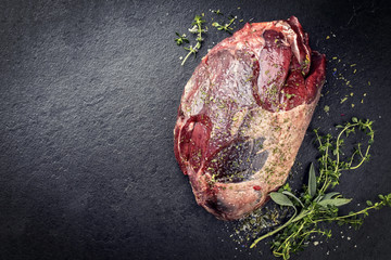 Raw dry aged haunch of venison as with herbs as close-up on a slate slab with copy space left