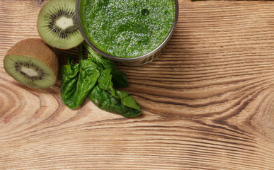Side of the corner on a wooden old table can be seen part of the glass, which is a green vitamin detox smoothies and next to the ingredients are ripe kiwi and juicy spinach leaves