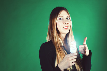 Fototapeta premium Serious young business woman holding a light bulb on a green background. Model shows a gesture Peace. Hands of a close-up girl
