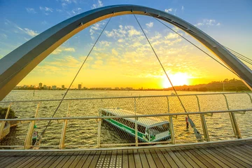 Poster Turistic ferry crossing iconic Elizabeth Quay Bridge at sunset on Swan River. Scenic landscape seen from wooden walkway of arched pedestrian bridge of Elizabeth Quay marina. Perth, Western Australia. © bennymarty