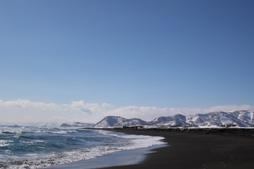Beautiful winter seascape of Pacific ocean with big waves, black volcano sand and snowy mountains background. View of Avacha bay on Kamchatka Peninsula nearby the city Petroplavlovsk-Kamchatsky.