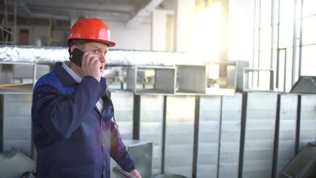 Young caucasian engineer talking on the phone at the building under construction. Architector in orange hard hat holding his phone near his ear. 4 k airduct of an HVAC system in building. Industrial