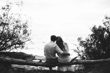 Romantic black and white photo, on which a couple in love sits on a log on the seashore, hugging and admire the scenery