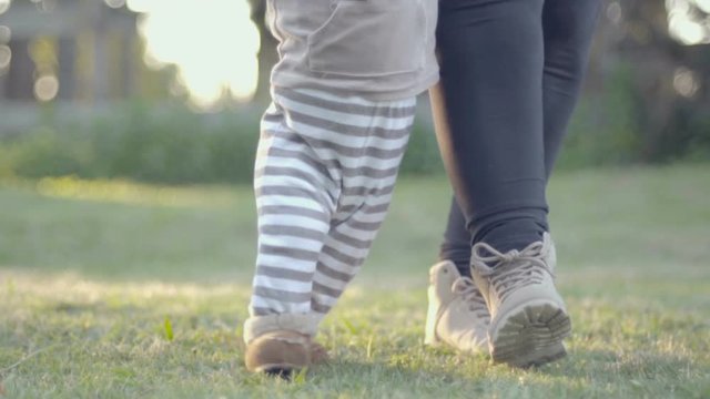 Baby boy walking first steps outdoor in the grass with mother help. Low angle view of toddler legs and feet.