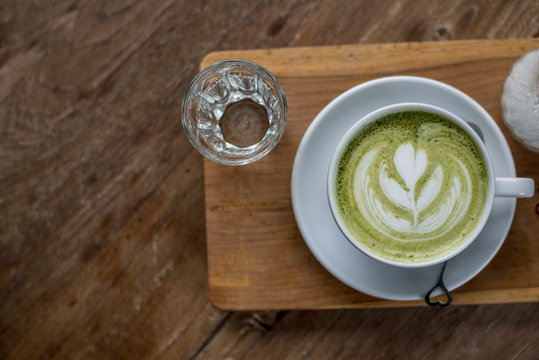 Hot Matcha Green Tea Latte in White Cup on Wood plate with some Water in Glass on Wooden table with some space on left side