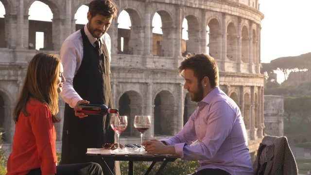 Elegant waiter serving pouring glass of red wine to romantic couple sitting at restaurant table in front of colosseum in rome at sunset
