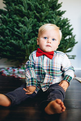 Naklejka premium Little boy sitting in front of the Christmas tree in a plaid shirt and red bow tie