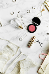Overhead view of female lace lingerie, perfume, lipstick, blushes and jewelry items on white marble background. Top view, text space. Beauty routine concept