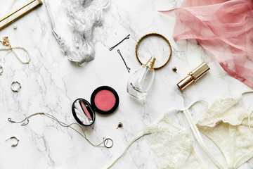 Overhead view of female lace lingerie, perfume, lipstick, blushes and jewelry items on white marble background. Top view, text space. Beauty routine concept