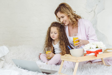 mother and daughter with glasses of orange juice using laptop in bed with breakfast on tray