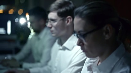 Rack focused shot of IT professionals working on computers in dark office at night with projector lightning at camera in background