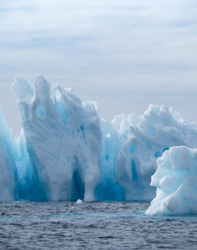 Iceberg with light and medium turquoise blues in a deep blue Southern Ocean. Cloudy sky is above. The icebergs have fissures, crevices and caves.
