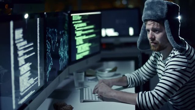 Tilt up of Russian hacker in fur ear flap hat and sailor shirt hacking information in office at night. Man working on computer with multiple monitors, projector lightning in background