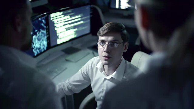 Tracking shot of young IT professional programmer in eyeglasses working on computer in cyber security center filled with display screens and talking with colleagues