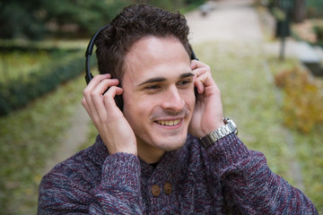 Cheerful young man standing and listening to music with headphones in autumn park. 