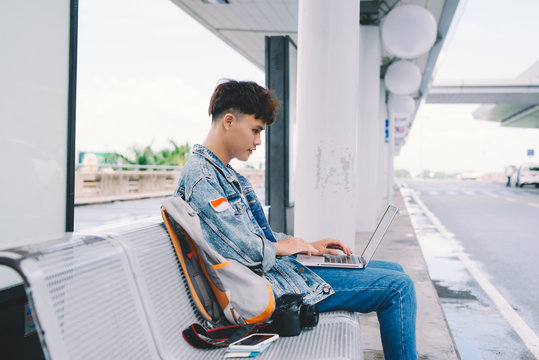 Asian young man sitting on the chair at the airport bus stop and using laptop