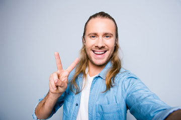 Close up picture of attractive, bearded blogger with long hair shooting selfie, gesturing peace symbol with two fingers, having life video chat,  standing over grey background