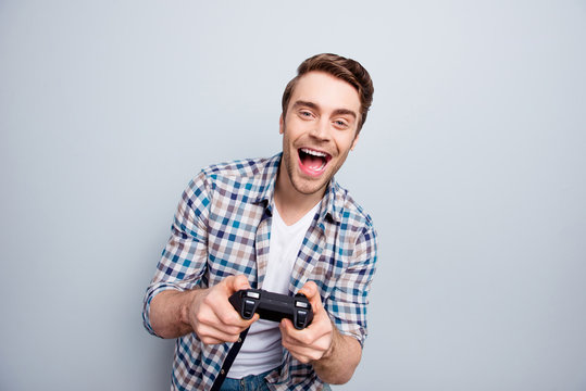Portrait of crazy, cheerful, attractive, positive, very excited guy in checkered shirt holding joystick and playing video games with wide open mouth over grey background