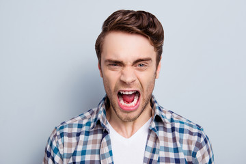 Aggressive man in checkered shirt with wide open mouth is out of himself, yelling, screaming, shouting with cruelty over grey background