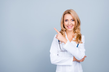 Look there! Portrait of confident experienced qualified cheerful smiling with blonde hair mature female doc, she is pointing on blank empty space over her shoulder, isolated on grey background