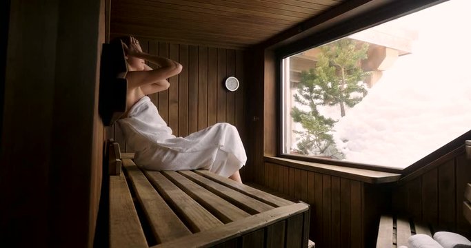 A beautiful woman wearing a white towel takes a sauna: The sauna is made of wood with a large window with a view of the snow. Concept of: relax, vacation, wellness center.