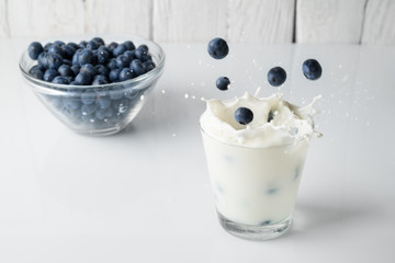 Fototapeta na wymiar Berries of blueberry fall into the glass with milk. Glass with milk and plate with berries on a white surface infront of a white wooden background.